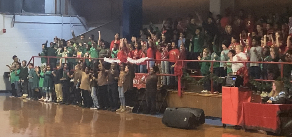 3rd and 4th grade students dressed in red, green, and brown shirts with jeans are standing in bleachers singing