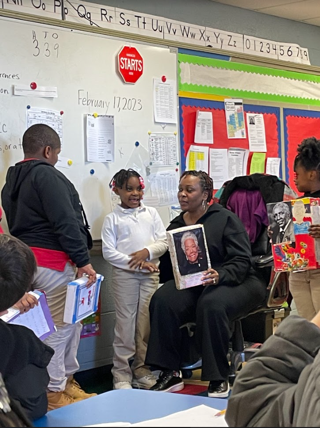 Ms. Gilland sitting with one student standing presenting her cereal box proejct