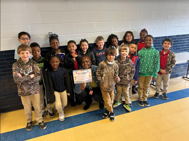 pictured are students standing against a wall in 2nd grade with their teacher who is holding a certificate